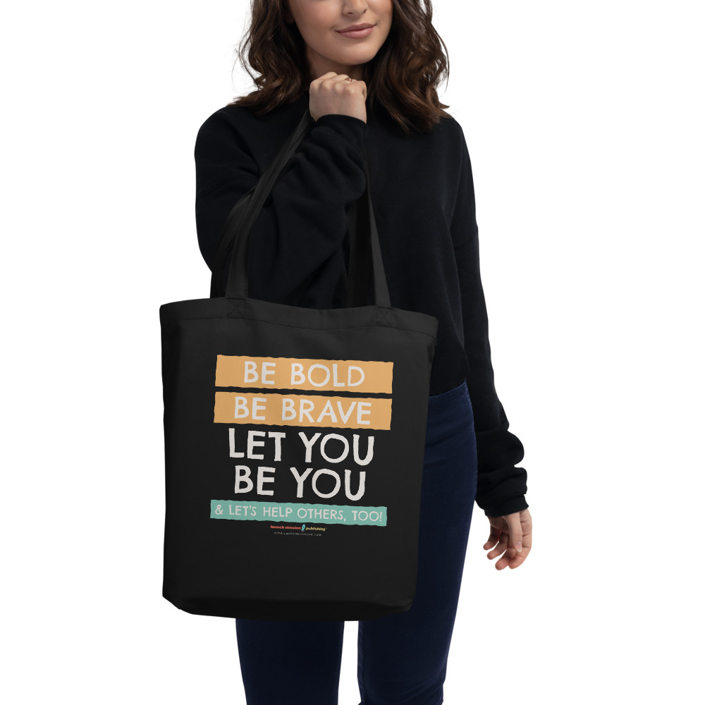 Adas-Motto-tote-bag_mockup_Front_with_model woman in all black clothes holding a black tote bag with the words "Be Bold, Be Brave, Let you be you. & Let's Help Others, Too!